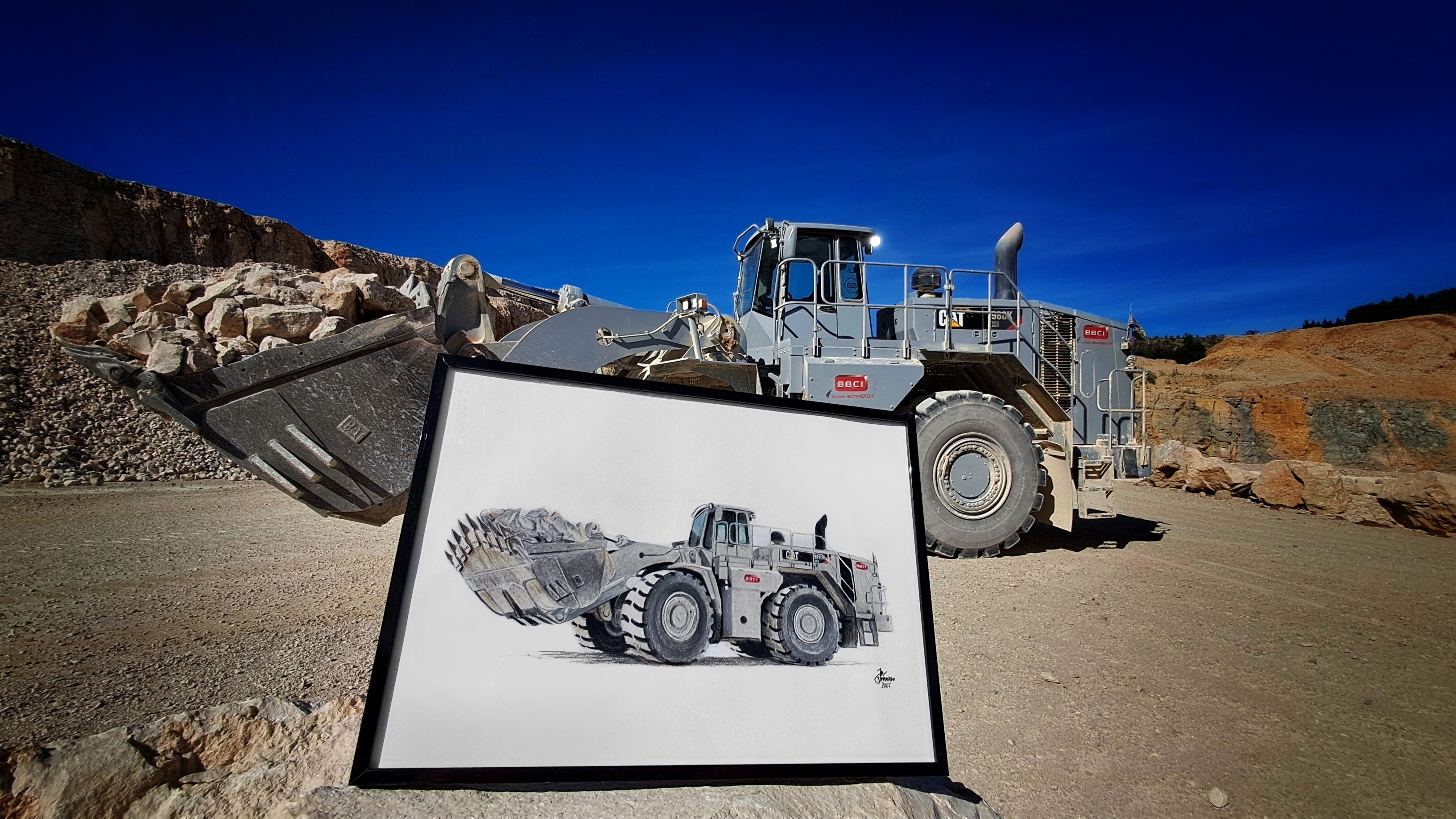 A framed pencil drawing of a CAT 988K XE wheel loader held up in front of the real machine at a quarry site